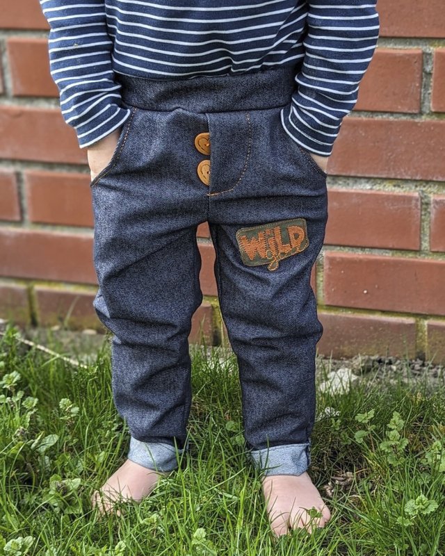 Kind trägt dunkelblaue Hose in Jeansoptik mit aufgenähtem orangefarbenem 'WILD'-Patch, zwei sichtbaren Holzknöpfen, Eingrifftaschen und umgeschlagenen Bündchen; barfuß auf Gras vor einer Backsteinmauer