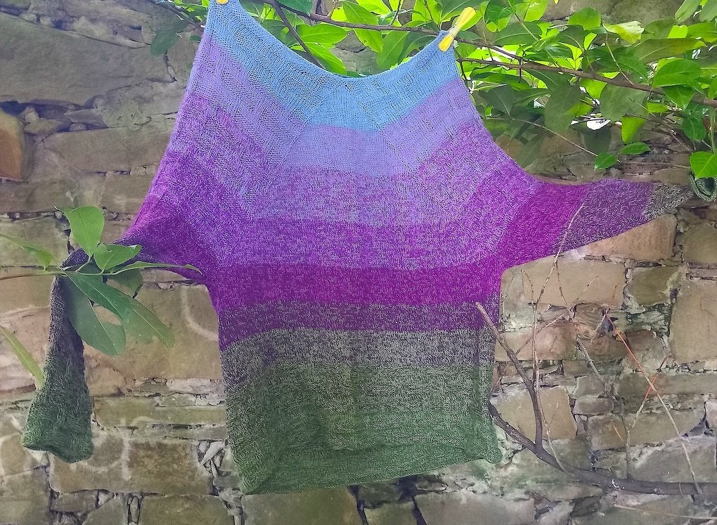 Weiter gestrickter Pullover mit Farbverlauf in Blau, Violett und Grün an einer Leine vor Steinmauer