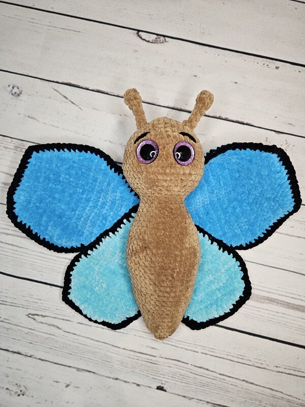Gehäkelter Amigurumi‑Schmetterling mit hellblauen und dunkelblauen Flügeln, braunem Körper und schwarzen Randkanten auf hellen Holzlatten