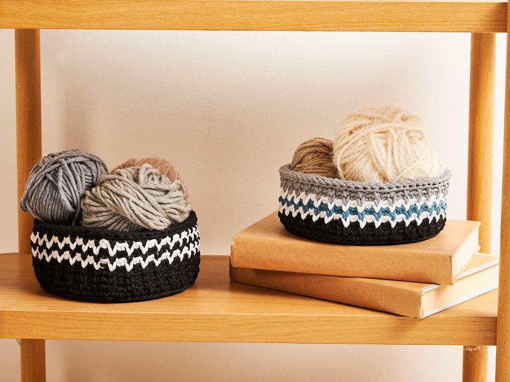 Zwei gehäkelte Körbe mit Zickzackmuster, gefüllt mit Wollknäueln, auf einem Holzregal