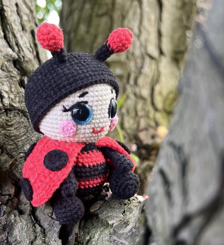 Red and black crocheted amigurumi ladybug doll with large glossy eyes sitting on tree bark