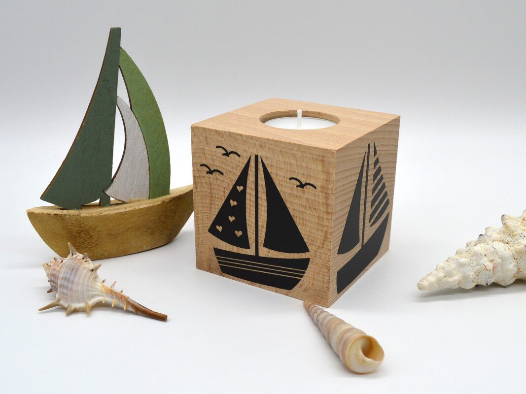 Quadratischer Holzteelichthalter mit schwarzem Segelbootmotiv, daneben eine kleine grüne Holzsegelbootfigur und Muscheln.