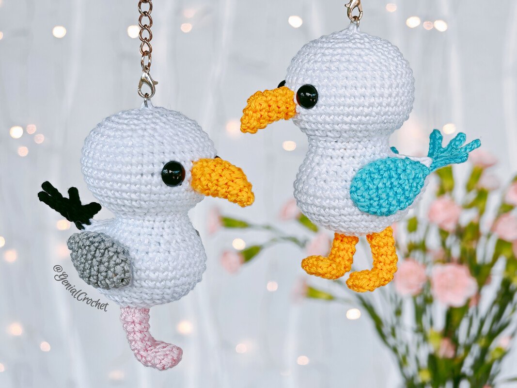 Two white crocheted bird keychains with yellow beaks and contrasting wings hanging on metal chains