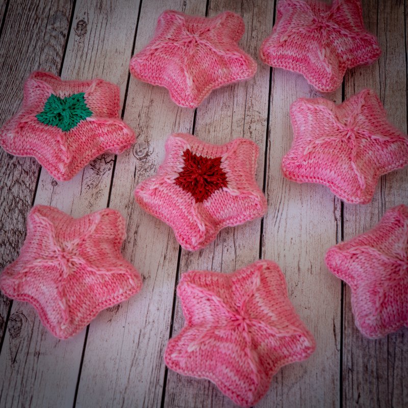 Gestrickte rosa Seesterne auf hellem Holz, zwei mit farbiger Mitte in Grün und Rot.
