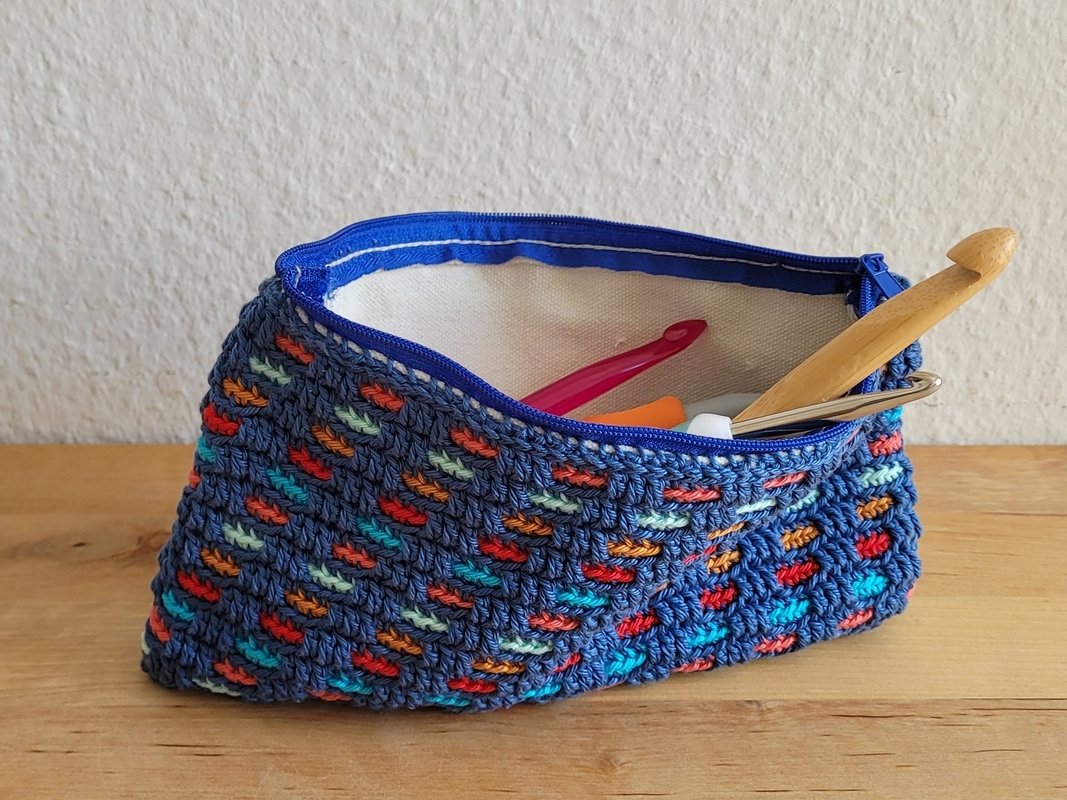 Gehäkelte dunkelblaue Reißverschlusstasche mit bunten Akzenten und offenem Innenraum, darin Häkelnadeln auf einem Holztisch
