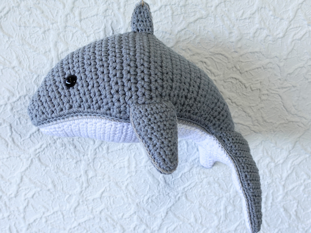 Gehäkelter grauer Amigurumi-Wal mit weißem Bauch vor strukturierter Wand