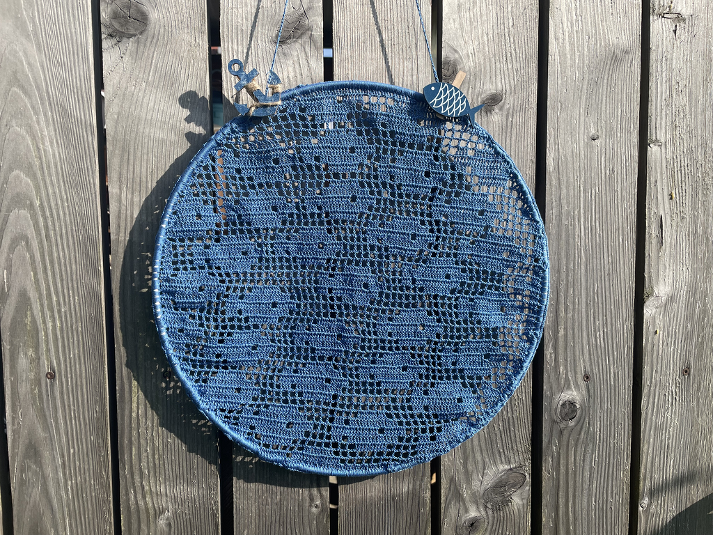 Rundes blaues Filethäkel-Wandbild in rundem Rahmen an einer Holzwand mit kleinen Holzanhängern