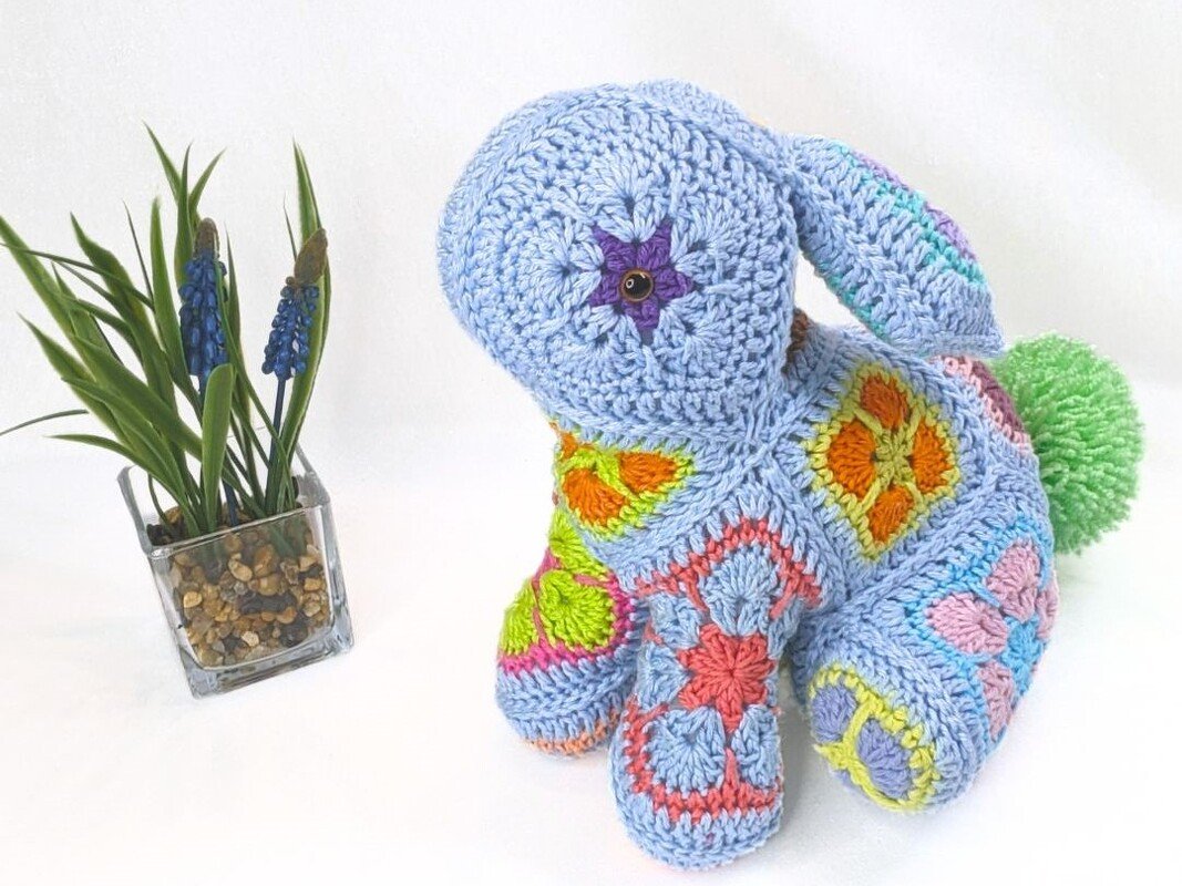 Light blue crocheted amigurumi bunny with colorful African Flower granny motifs, button eye, and green pom-pom tail beside a small potted plant.