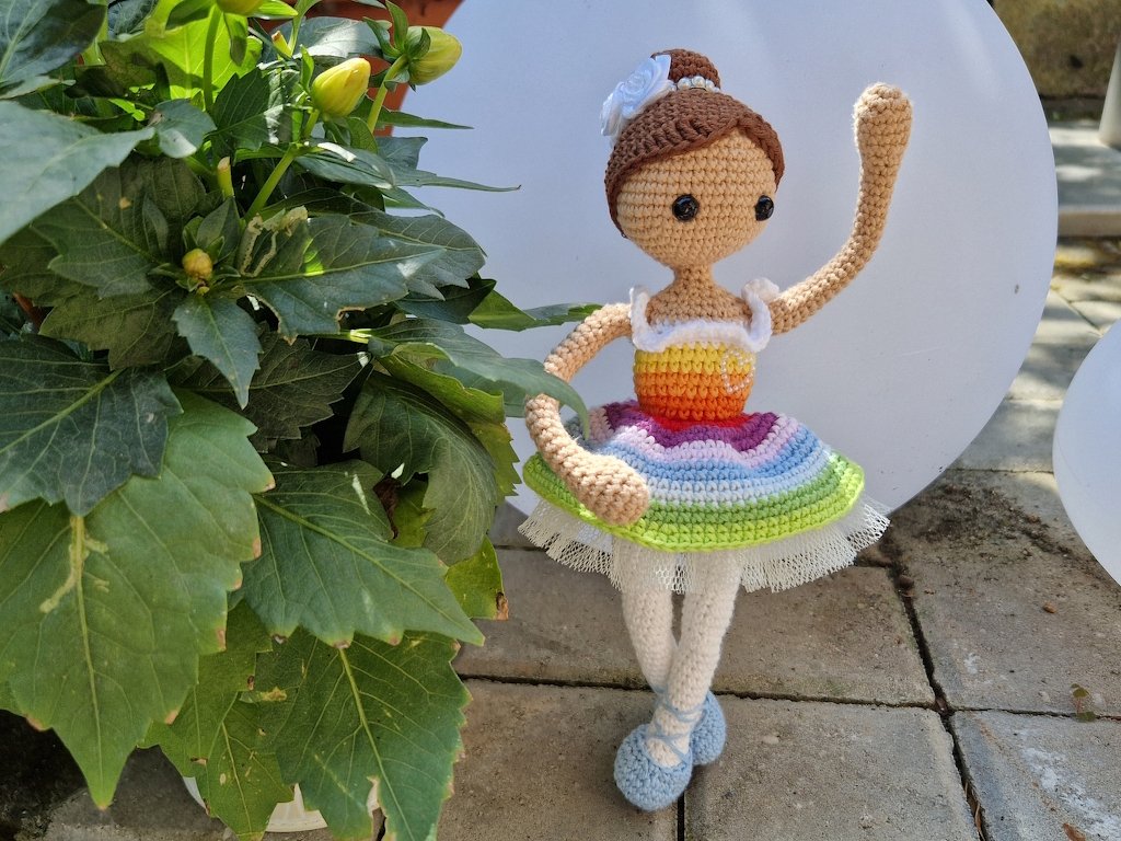 Gehäkelte Amigurumi-Puppe als Ballerina mit Regenbogenrock, weißer Blüte im Haar, weißen Beinen und blauen Schuhen