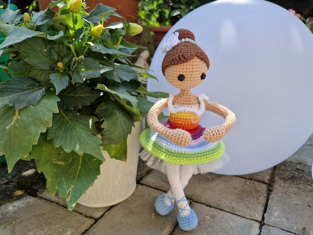 Gehäkelte Amigurumi-Ballerina mit Dutt, Regenbogen-Tutu und hellblauen Ballettschuhen auf Terrasse