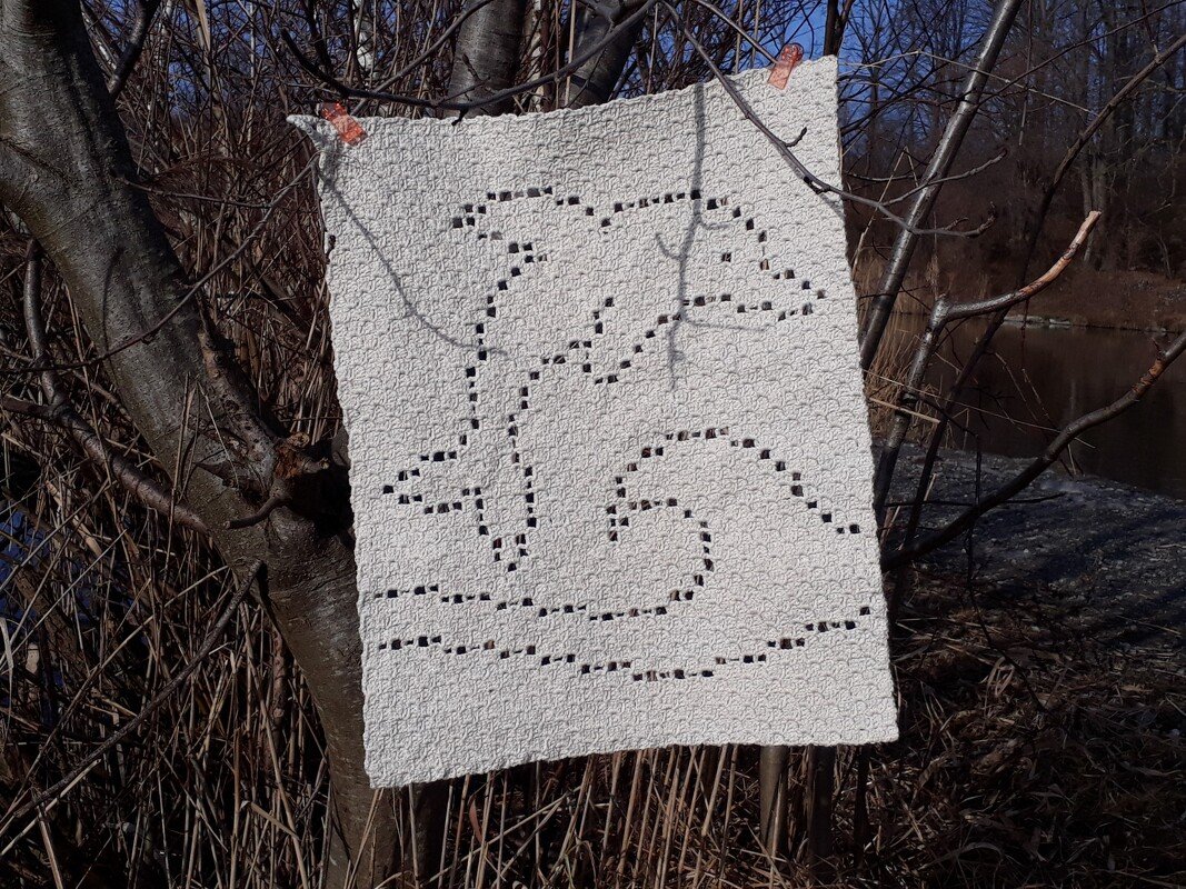White filet crochet square showing two jumping dolphins over a wave, clipped to tree branches