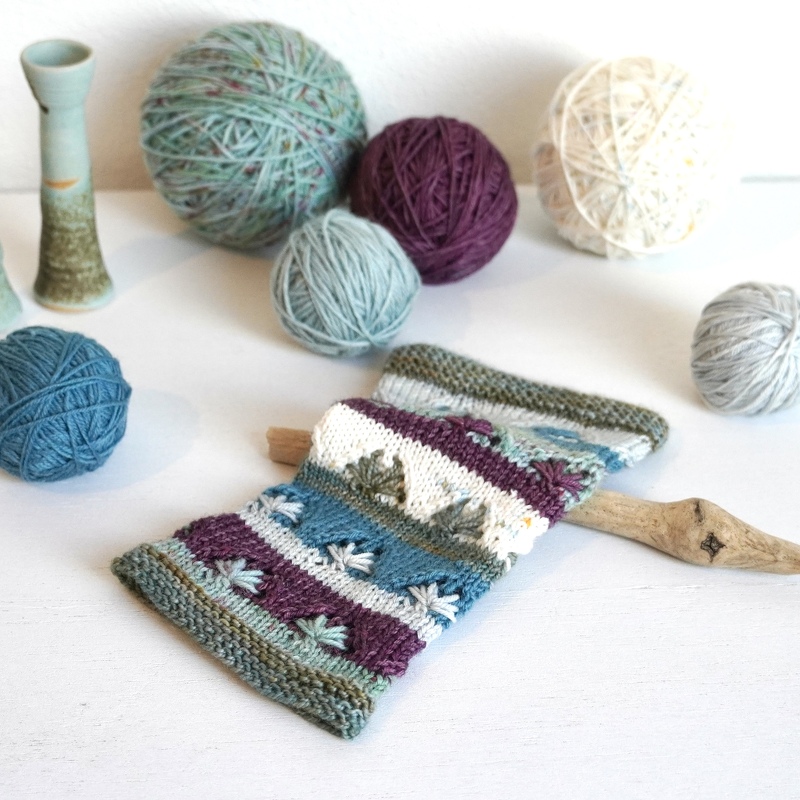 Nahaufnahme handgestrickter Armstulpen in Lila, Türkis und Grün mit Blattmotiv, umgeben von Wollknäueln.