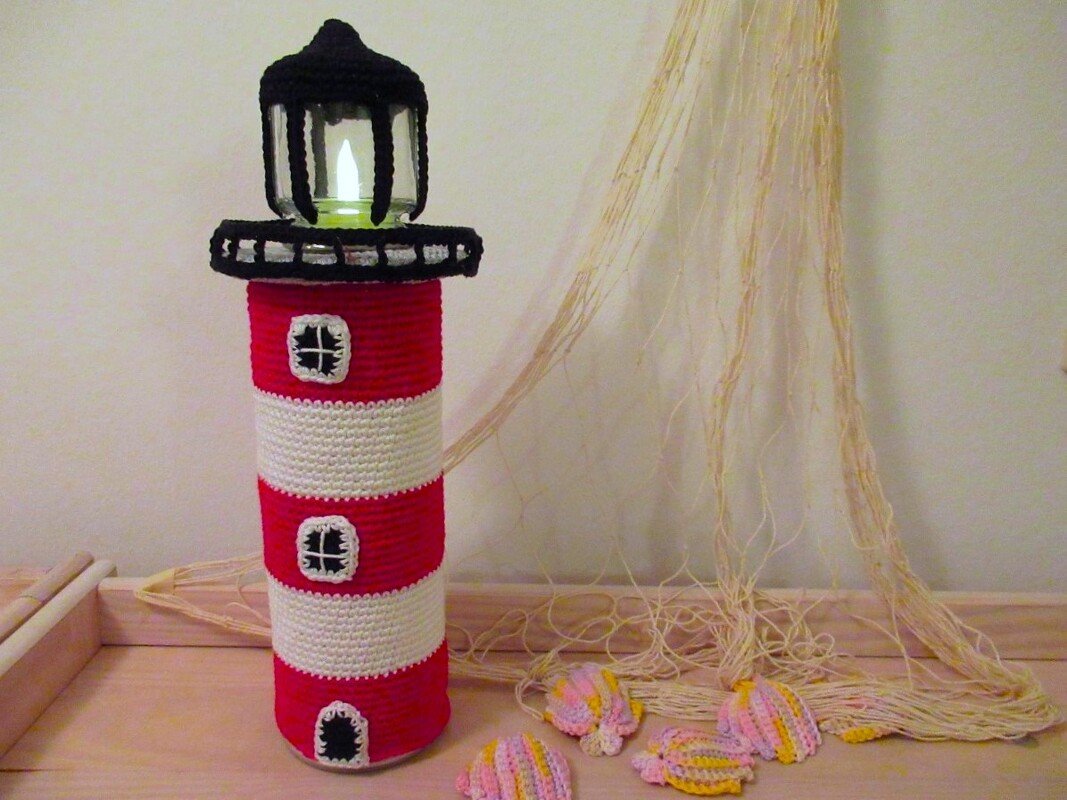Handgehäkelter rot-weißer Leuchtturm mit schwarzer Laterne und LED-Kerze, neben Fischernetz und gehäkelten Muscheln.
