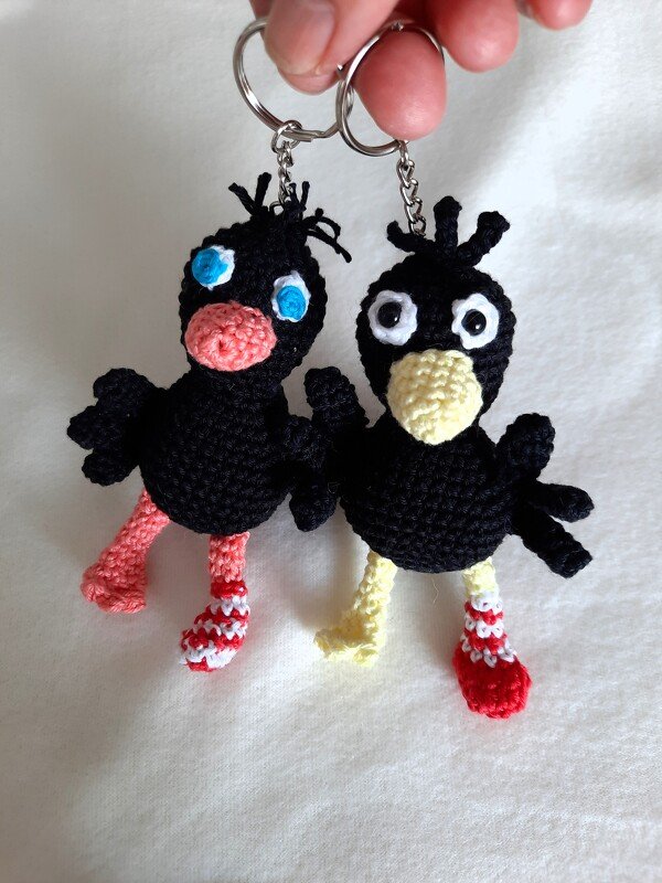 Zwei gehäkelte schwarze Vogel‑Amigurumi als Schlüsselanhänger mit bunten Schnäbeln und gestreiften Socken an Metallringen