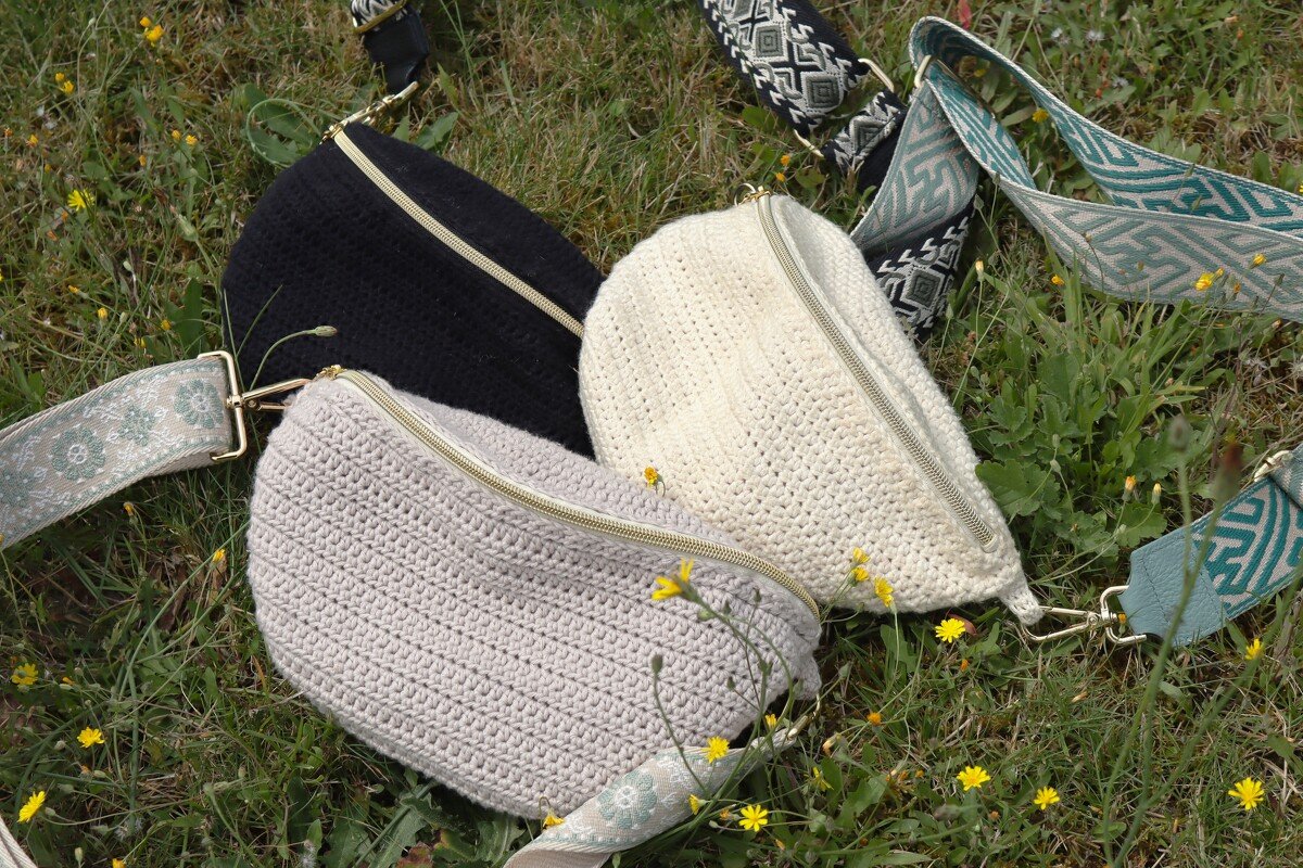Three crocheted sling bags in black, off-white and light grey with wide patterned fabric straps and gold zippers on grass.