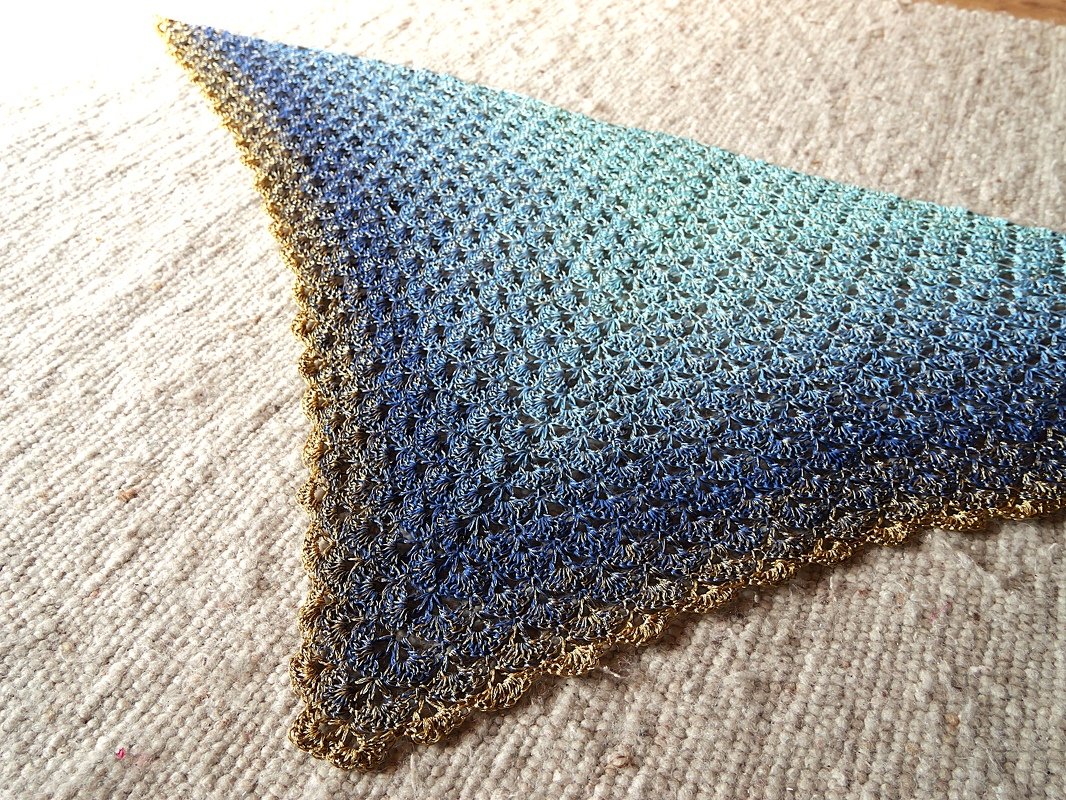 Dreieckiges gehäkeltes Tuch mit blau-türkis Farbverlauf und goldfarbener Muschelkante auf hellem Teppich