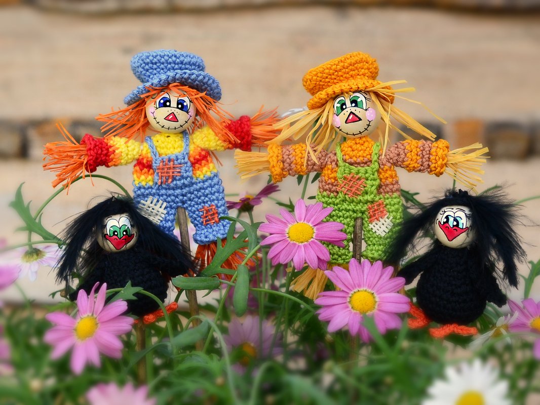 Zwei gehäkelte Vogelscheuchen mit Raffia-Haar und zwei schwarze Häkelraben zwischen rosa Blumen