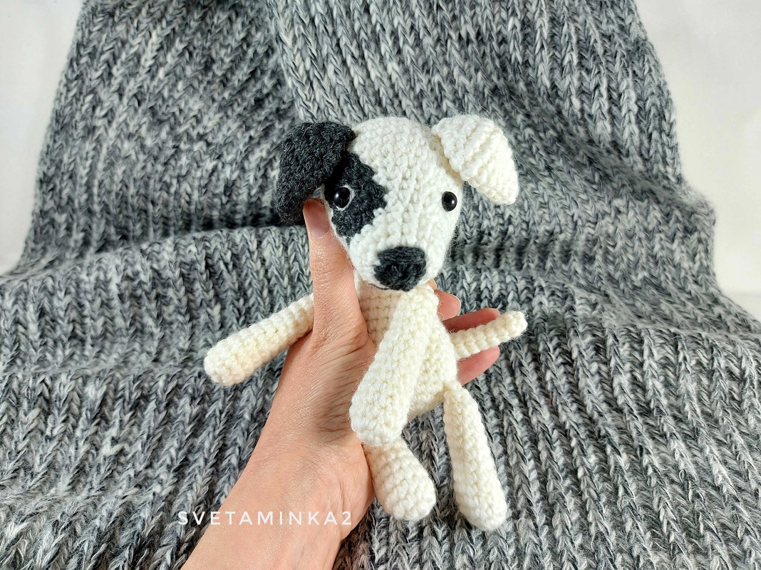Hand holding small white crochet amigurumi dog with black eye patch and black snout on grey knit blanket