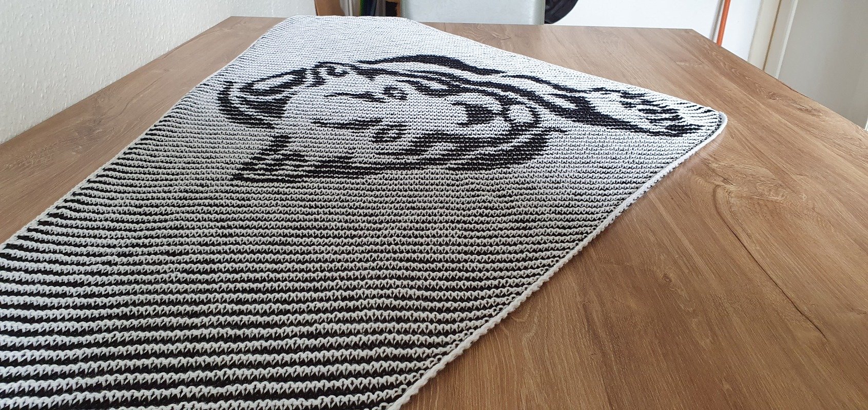 Black and white knitted shawl with a stylized creature motif on one end, laid flat on a wooden table.