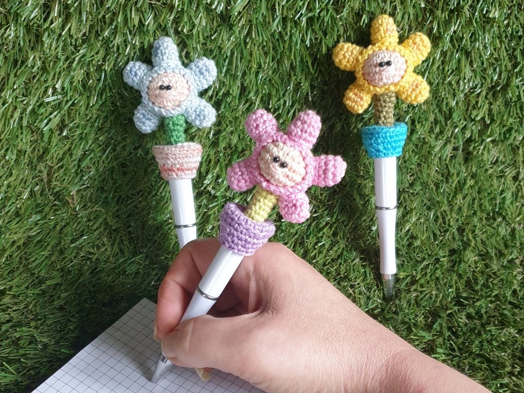 Nahaufnahme von drei weißen Stiften mit gehäkelten Blumenaufsätzen in Pastellfarben; eine Hand hält den mittleren Stift über kariertem Papier