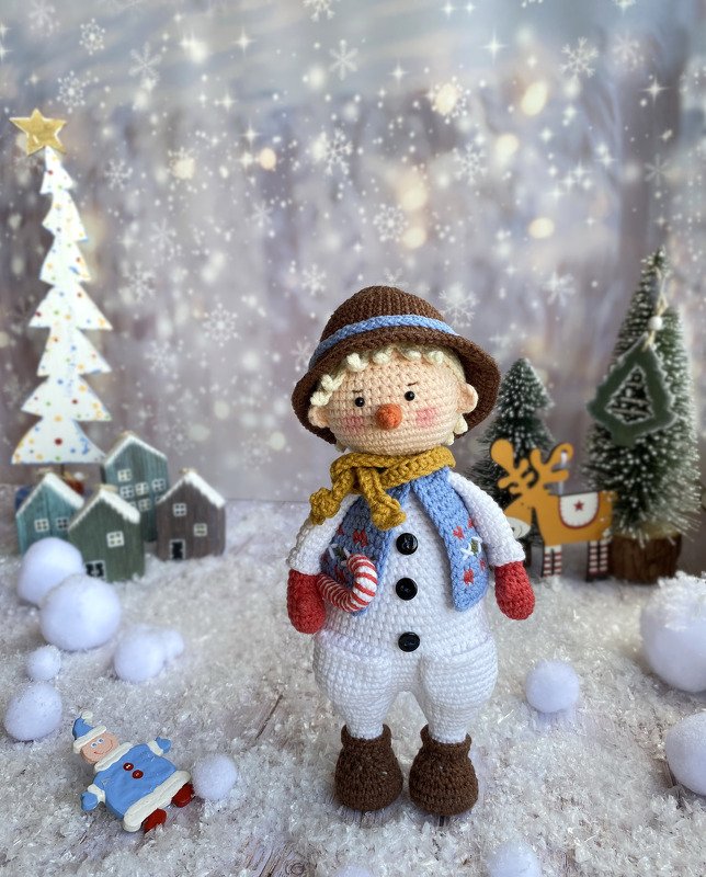 Gehäkelter Amigurumi-Schneemann mit braunem Hut, gelbem Schal, blauer Weste und Zuckerstange in einer verschneiten Miniaturszenerie mit Tannenbäumen.