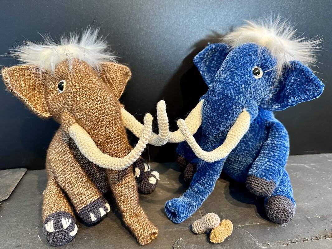 Zwei gehäkelte Amigurumi-Elefanten, einer braun, einer blau, mit hellen Stoßzähnen und weißer Mähne