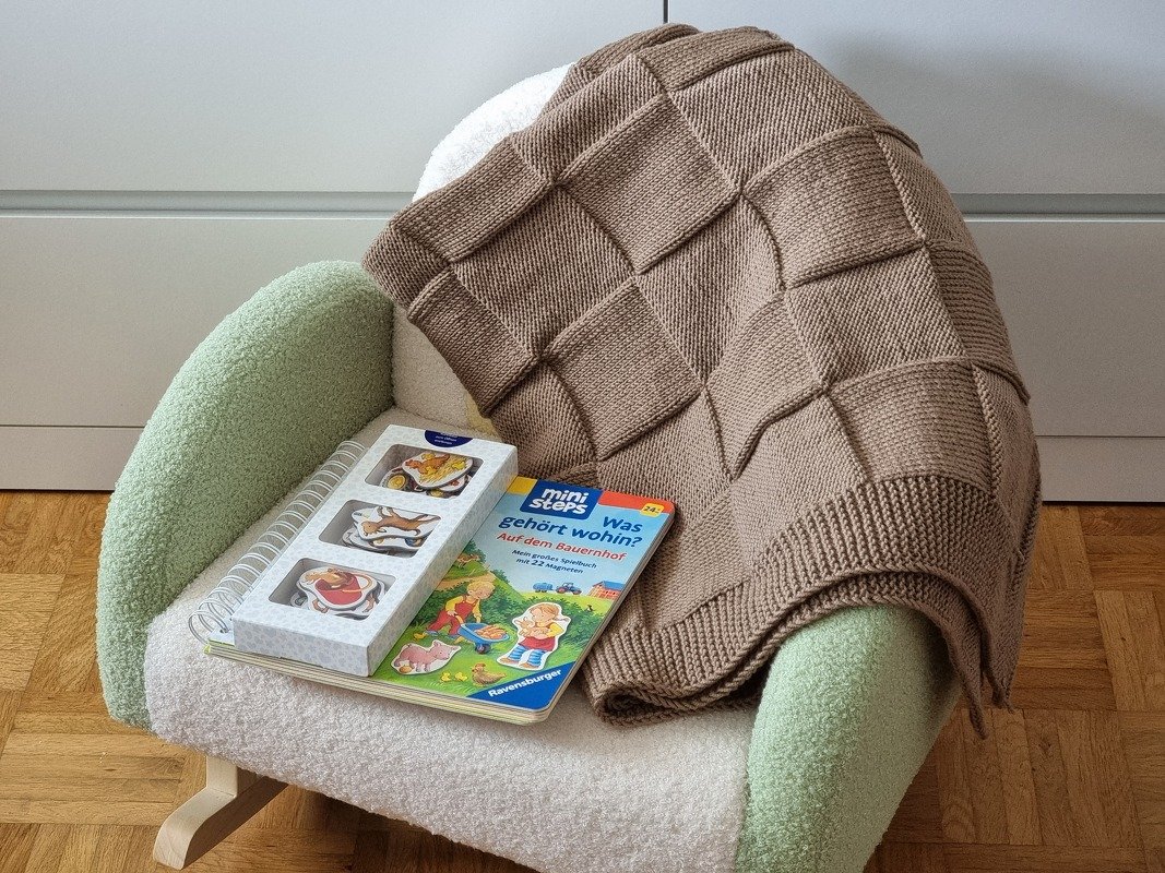 Braune, gestrickte Babydecke mit Relief-Karomuster auf einem Sessel neben Bilderbuch und Spielzeug.
