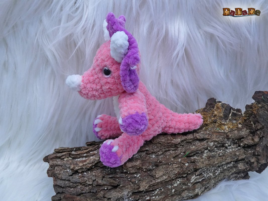 Gehäkeltes rosa-lila Amigurumi-Fantasiewesen auf Baumrinde vor weißem Kunstfell