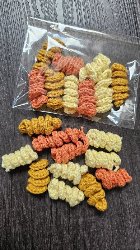 Mehrere gehäkelte Spiralnudeln in Gelb, Koralle und Creme, teils in einem durchsichtigen Plastikbeutel auf dunklem Holzuntergrund