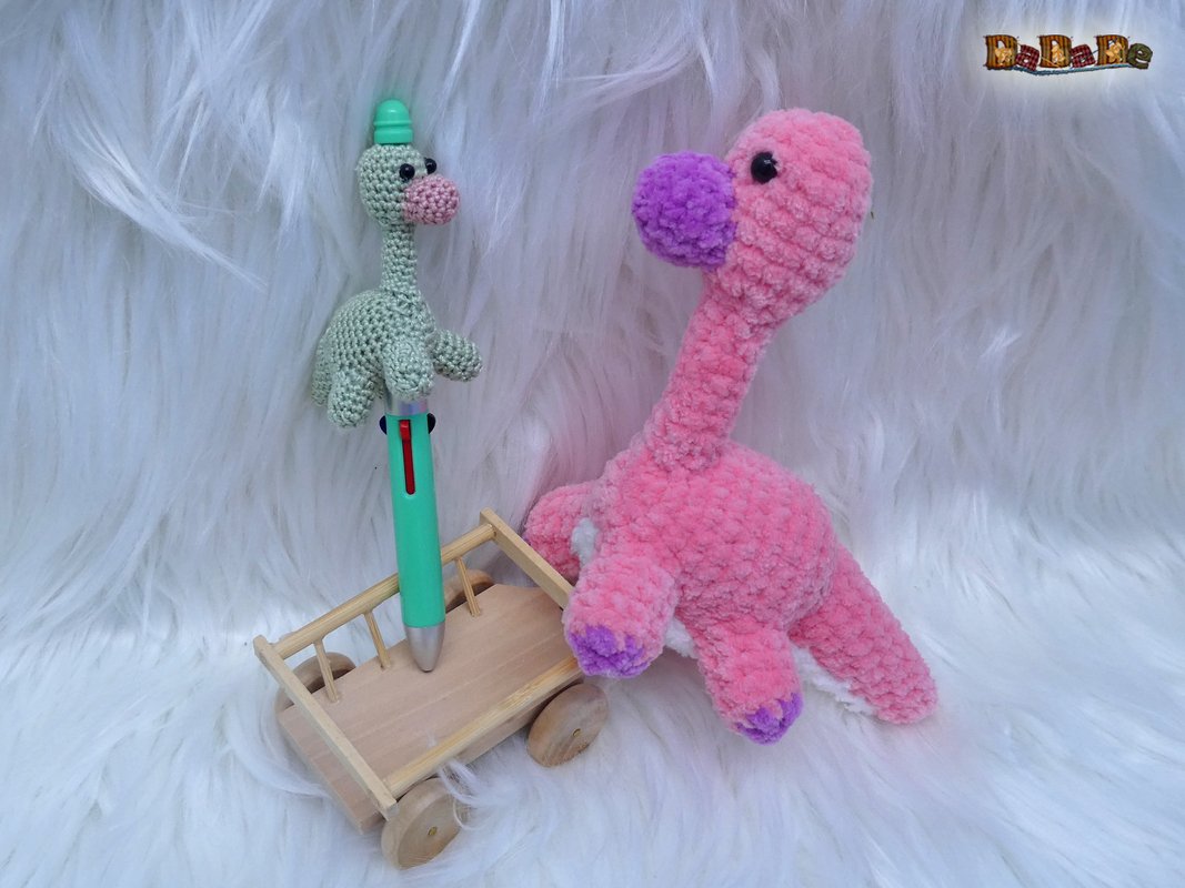 Pastellrosa flauschiger Dino-Amigurumi mit violetter Schnauze und Pfoten neben kleinem grünen gehäkelten Stiftaufsatz auf einem Holzwagen vor weißem Fell