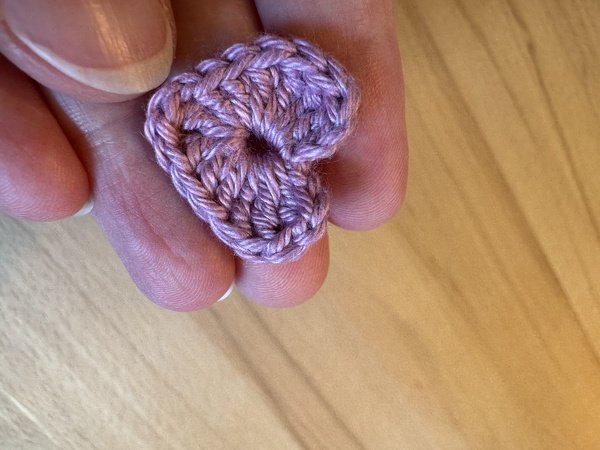 Hand hält ein kleines pastellfarbenes gehäkeltes Herz vor hellem Holzuntergrund