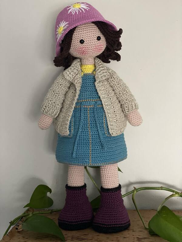 Front-facing crocheted amigurumi doll with pink daisy hat, cream cardigan, blue dress and purple boots