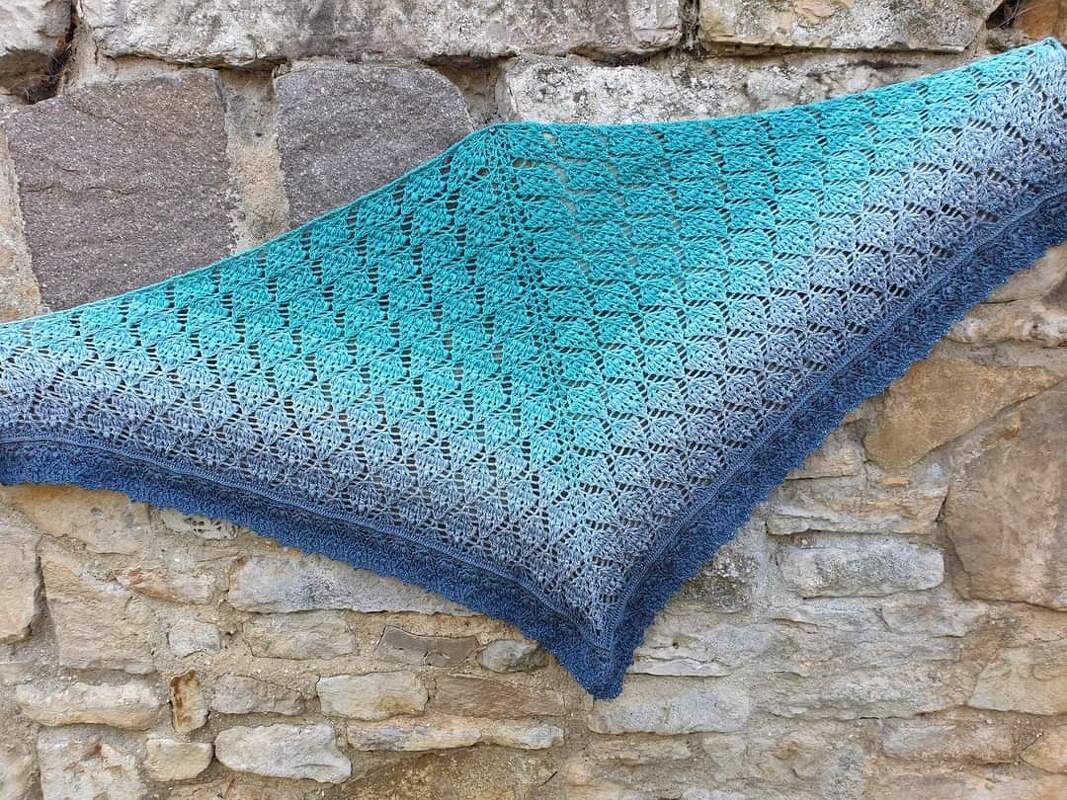 Gehäkeltes Dreieckstuch in Blau-Türkis mit Ajourmuster vor einer Natursteinwand