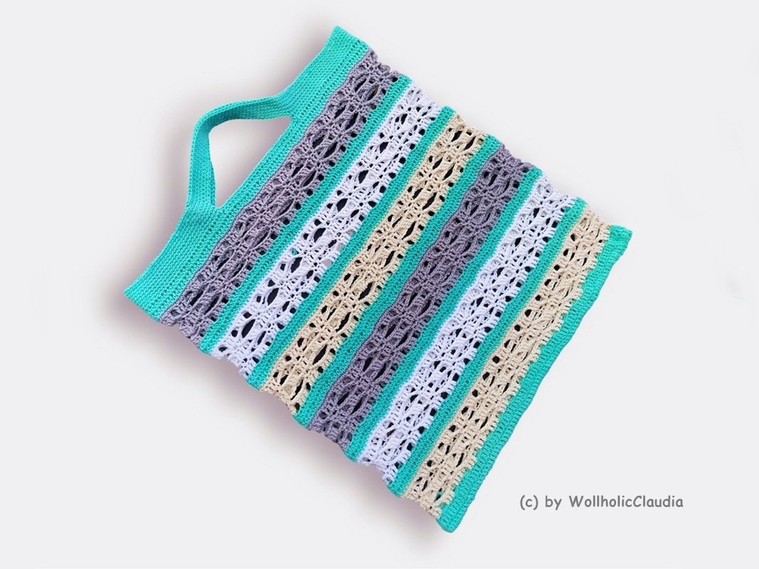 Flach liegender gehäkelter Shopper mit vertikalen Streifen in Türkis, Grau, Weiß und Beige; Henkel am oberen Rand sichtbar.