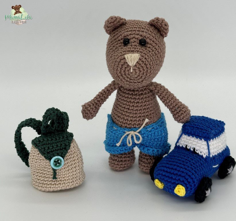 Brauner Amigurumi-Häkelbär mit blauen Shorts, daneben ein gehäkelter Rucksack und ein blaues Häkelauto