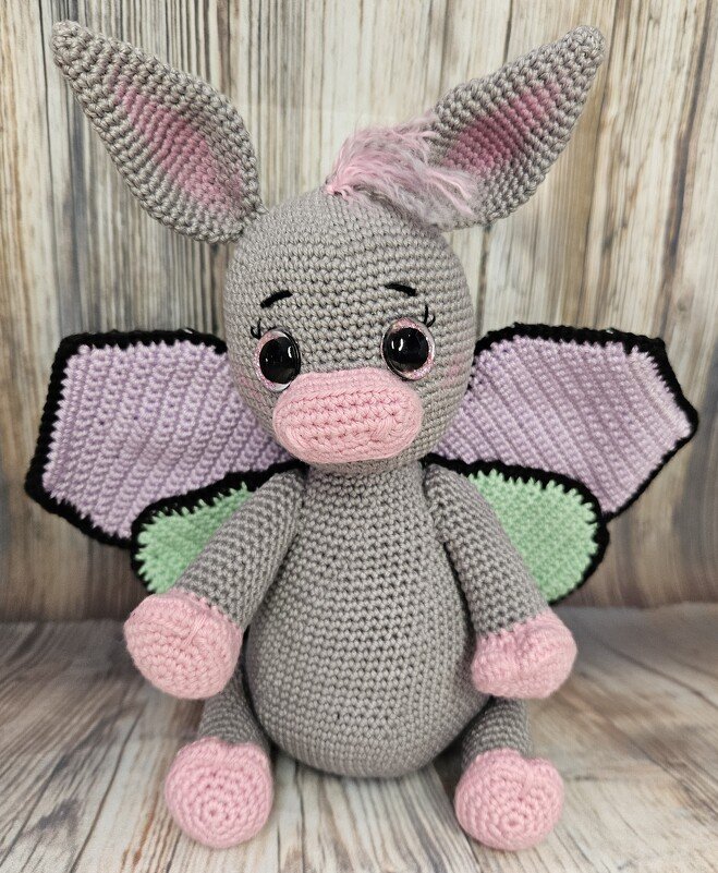 Gehäkelter grauer Esel mit rosa Schnauze, Pfoten und Ohren sowie lila-grünen Schmetterlingsflügeln.