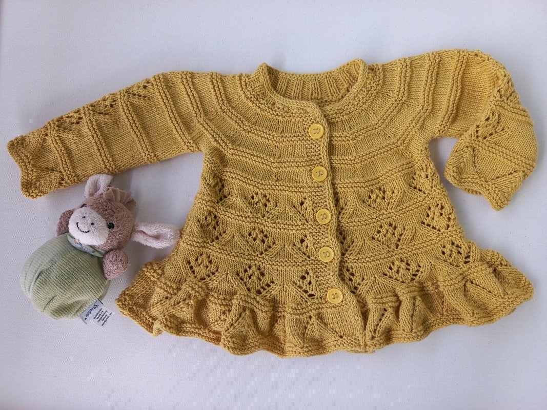 Senfgelbe gestrickte Babyjacke mit Lochmuster, fünf Knöpfen und Rüschensaum, daneben ein kleines Kuscheltier