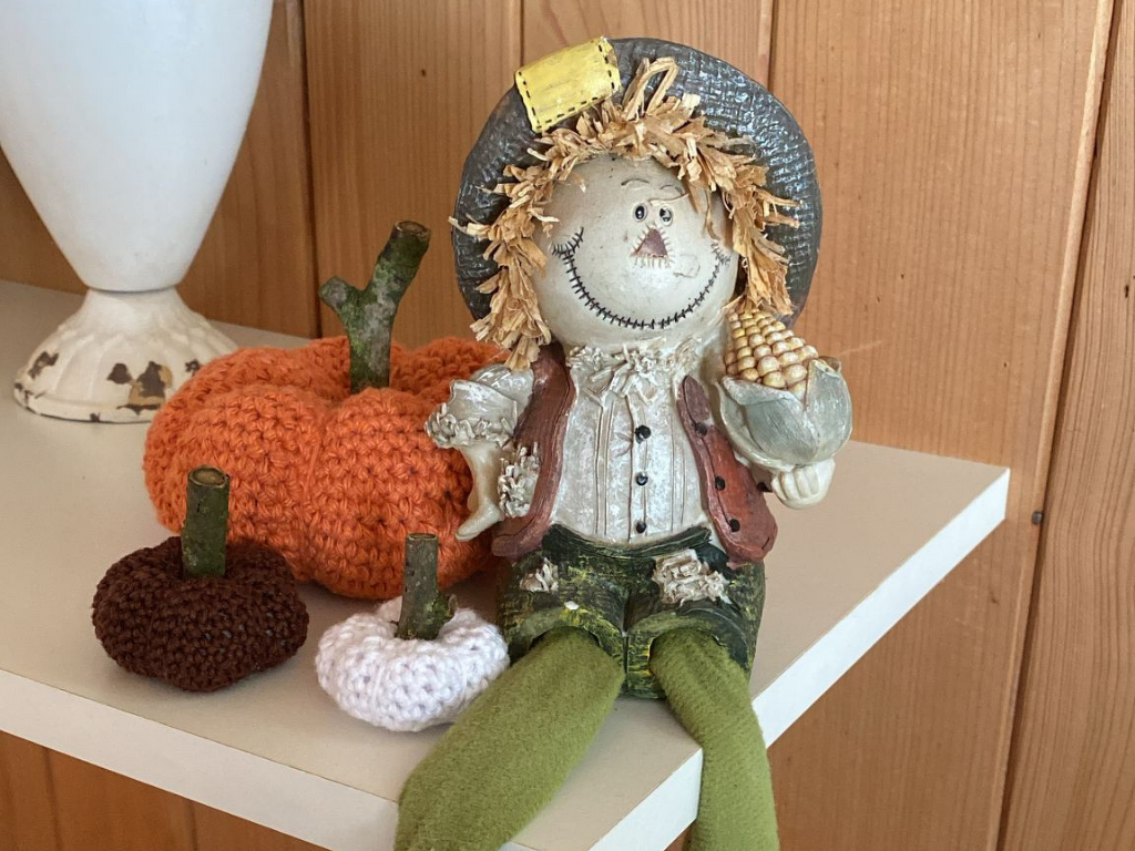 Herbstliche Deko mit gehäkeltem Kürbis-Set in Orange, Braun und Weiß auf einem Regal neben einer Vogelscheuche