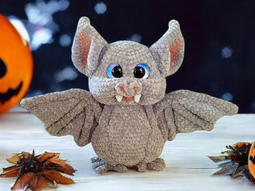 Gray crocheted bat amigurumi with iridescent blue eyes and small fangs, on a light wooden surface with Halloween decorations.