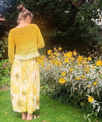 Person von hinten in gelbem Häkelpullover über hellgelbem, gemustertem Maxikleid vor blühenden gelben Blumen