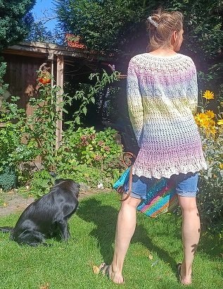 Rückenansicht einer Person in einem pastellfarbenen Häkelkleid mit Lochmuster im Garten neben einem schwarzen Hund.