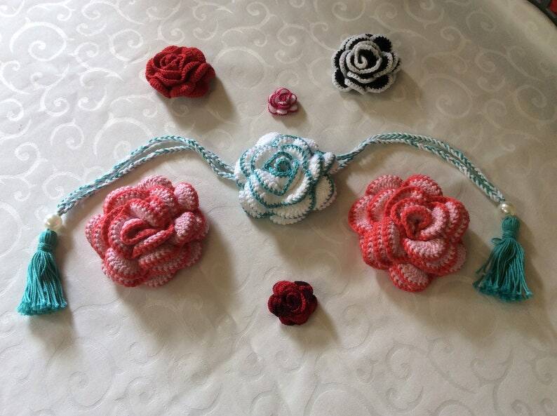 Assorted crocheted roses in red, pink, white, teal, and black on patterned fabric, with a braided garland and tassels.