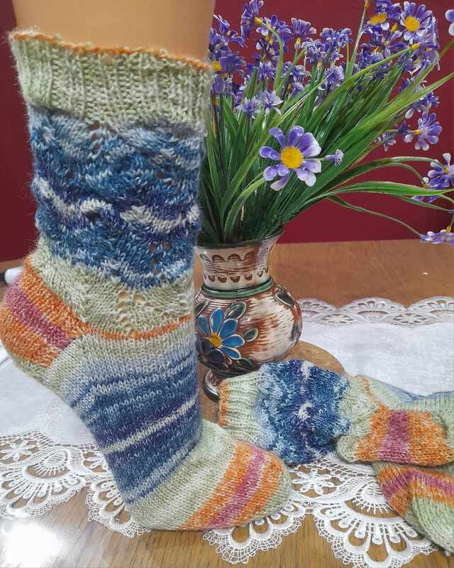 Handgestrickte Damensocken: eine am Fußmodell getragen, eine danebenliegend; Schaft mit blauem Lochmuster und mehrfarbigen Streifen, Vase mit lila Blumen im Hintergrund.