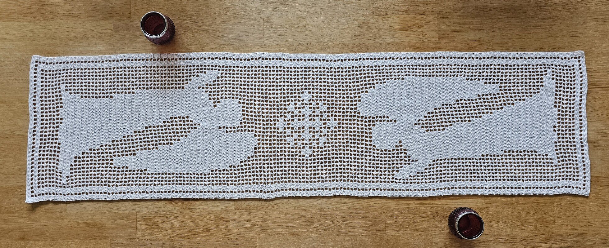 Weißer, handgehäkelter Tischläufer in Filethäkeltechnik mit Rentier- und Sternmotiven auf Holztisch, daneben zwei Kerzengläser.