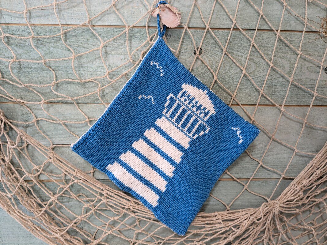 Blue-and-white knitted square potholder with a white lighthouse motif hanging on a fishing net against a light blue wooden wall.