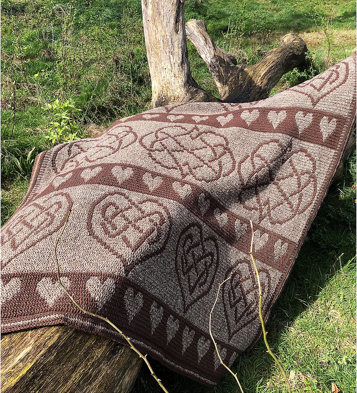 Brown and beige crocheted blanket with Celtic knot heart motifs draped over a log in a grassy outdoor setting.