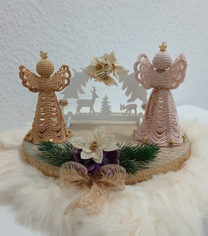 Zwei gehäkelte Engel (goldfarben und rosa) auf einem dekorierten Holztablett mit Tannenzweigen, Blumen und einer weißen Holzdeko mit Hirschmotiven, auf Kunstfell platziert.