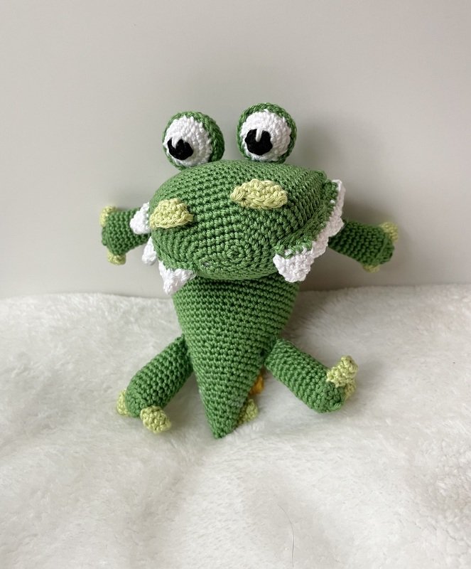 Gehäkeltes grünes Amigurumi‑Monster mit großen Augen und weißen Zähnen auf hellem Stoff
