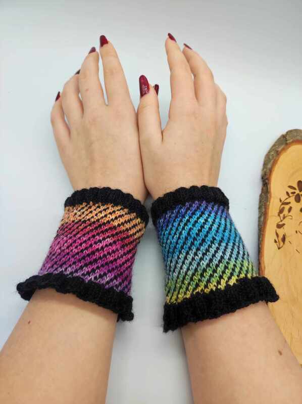 Hände mit bunten, diagonal gestrickten Armstulpen in Regenbogenfarben und schwarzem Rüschenrand