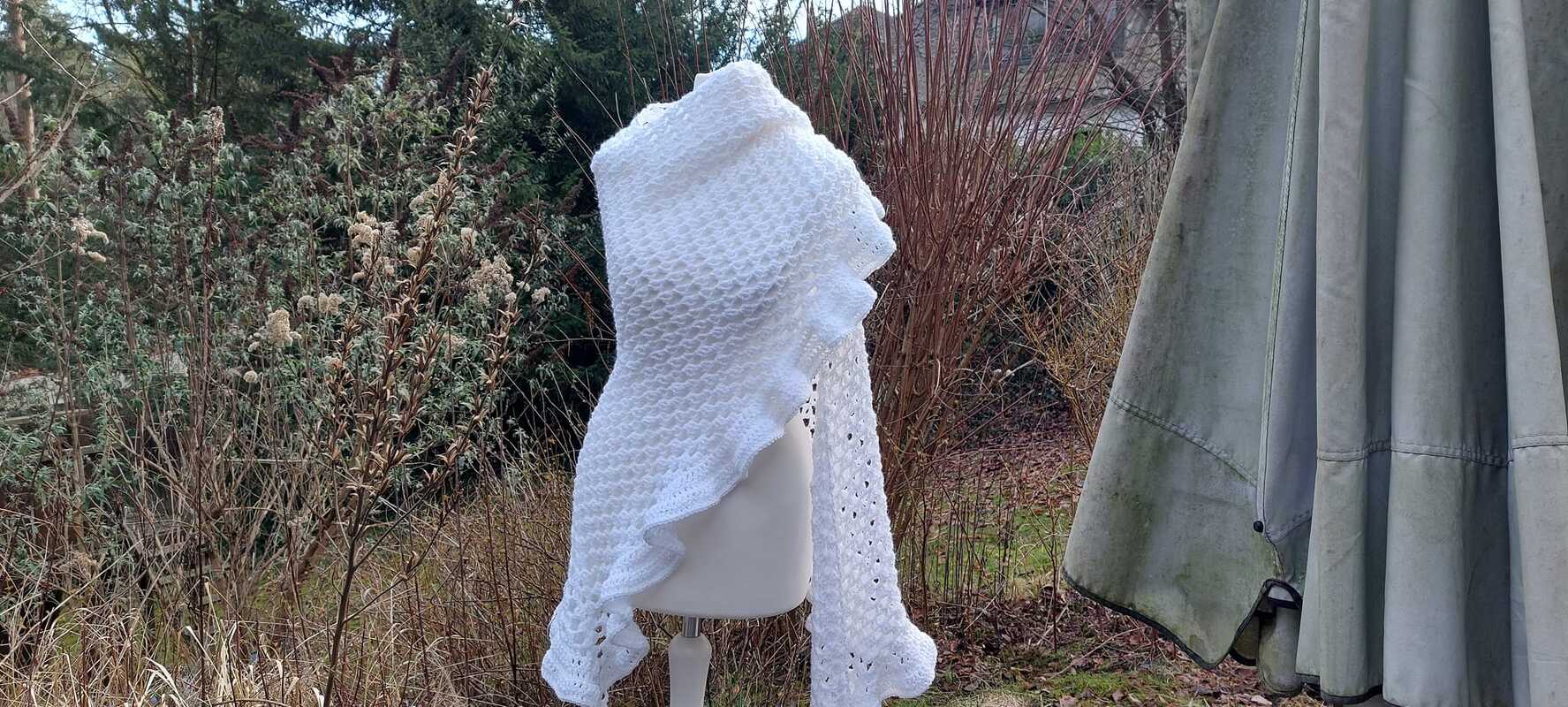 weißes gehäkeltes Dreieckstuch mit Rüschen auf Schaufensterpuppe vor winterlichem Garten